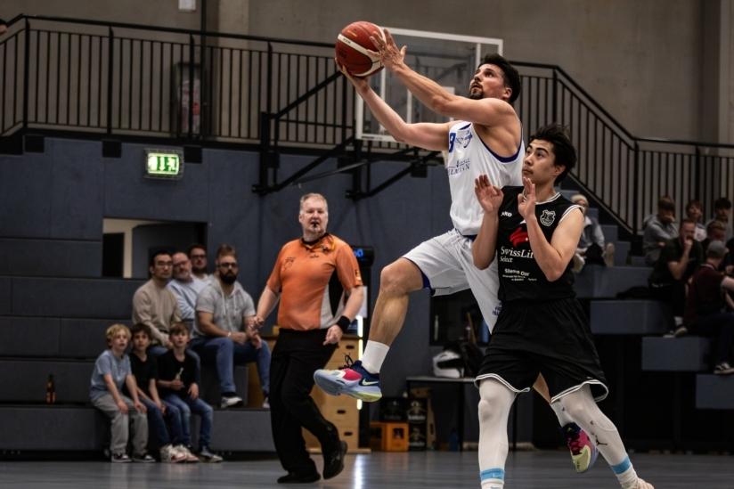 Basketballspieler springt für einen Korbwurf, während ein Gegner ihn verteidigt. Zuschauer sitzen im Hintergrund.