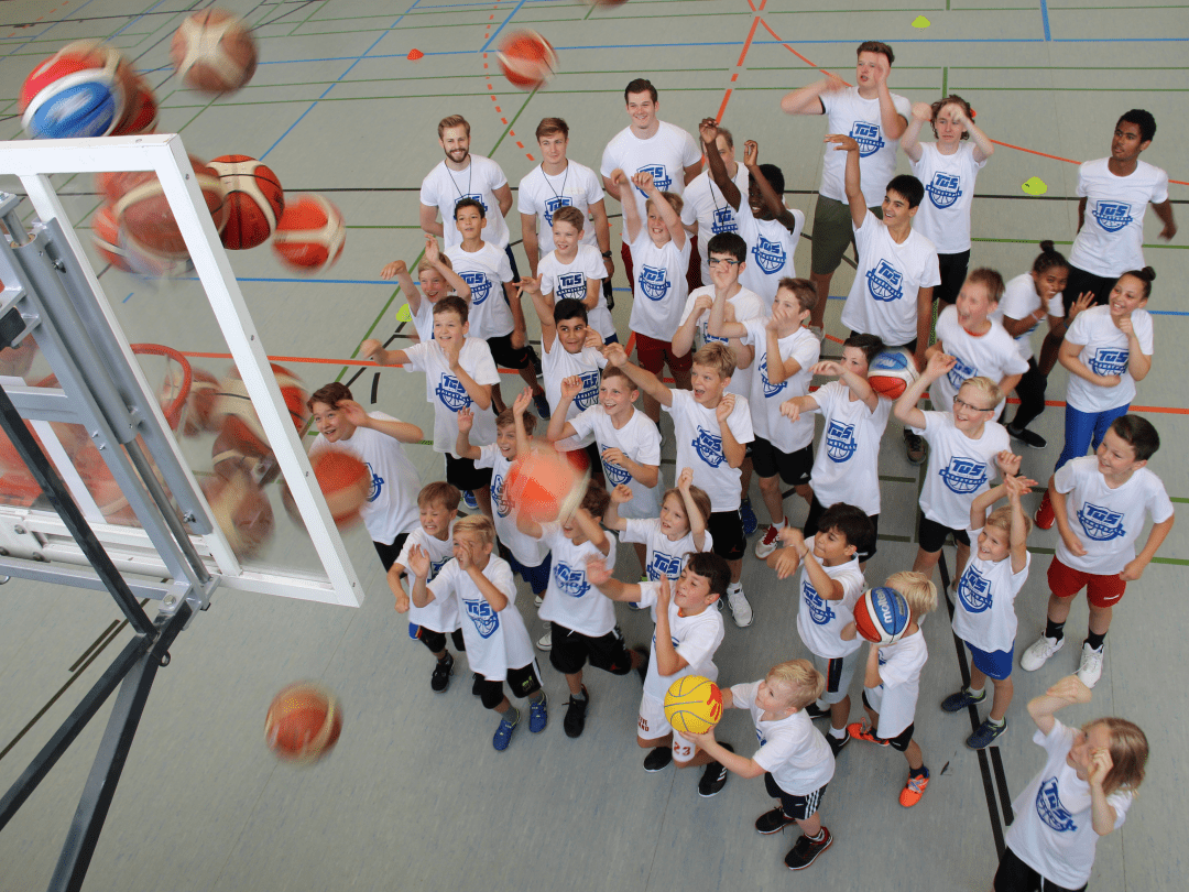 Gruppe von Kindern in weißen T-Shirts, die auf einen Basketballkorb zielen, während Bälle in der Luft fliegen.