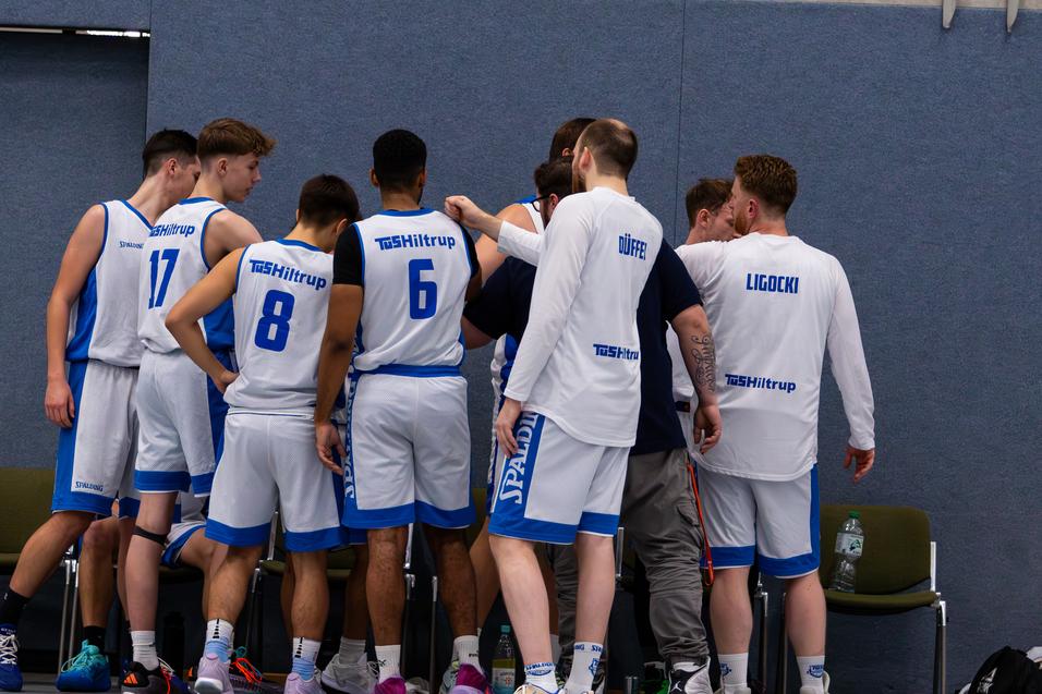 Mehrere Basketballspieler stehen in einem Huddle und besprechen strategie, während auf der Bank im Hintergrund Flaschen stehen.