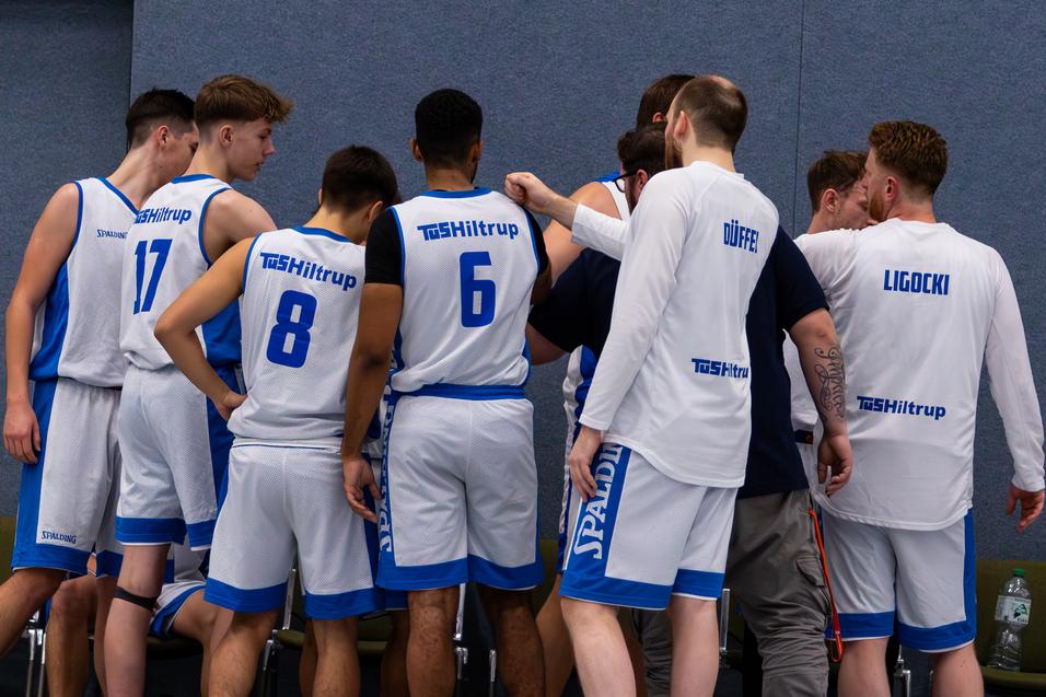 Mehrere Basketballspieler stehen in einem Huddle und besprechen strategie, während auf der Bank im Hintergrund Flaschen stehen.