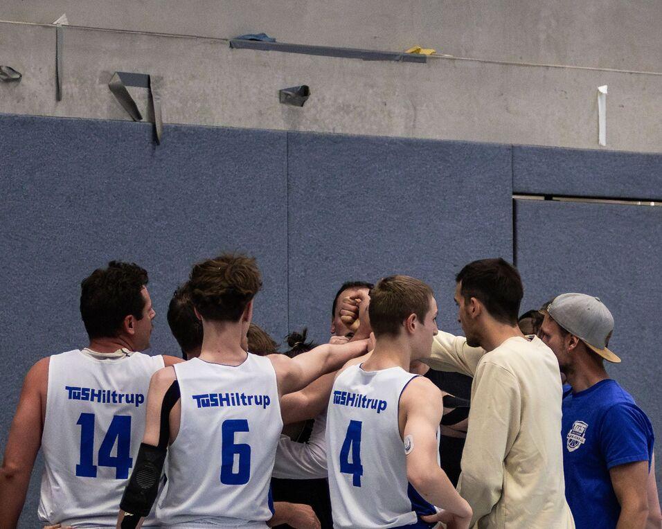Basketballmannschaft in weißer Sportkleidung berät sich im Kreis, während ein Spieler auf der Bank sitzt.