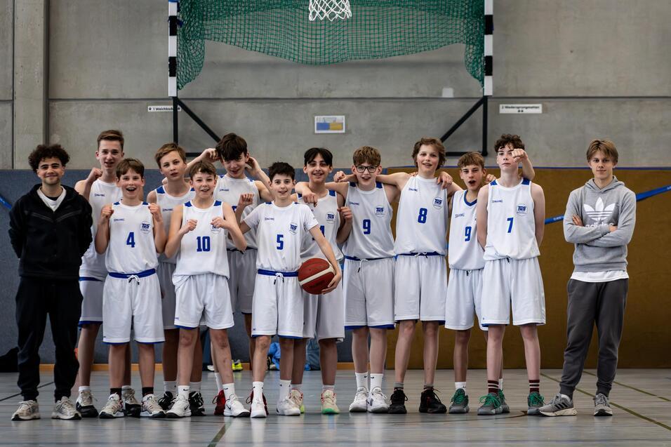 Gruppe von zwölf Jugendlichen in Basketballtrikots, posierend auf einem Sportplatz mit einem Basketball.