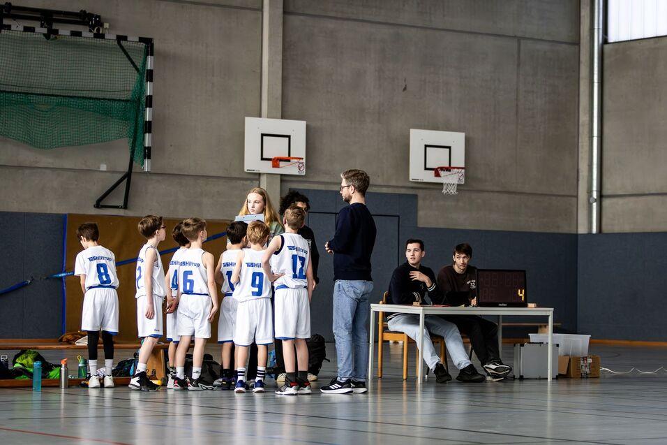 Gruppe junger Basketballspieler in weißer Sportkleidung versammelt sich um einen Coach in einer Sporthalle.