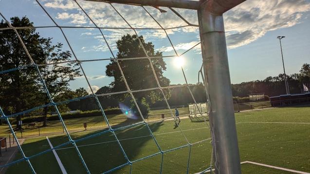 Blick durch ein Fußballtor auf einen Spieler, der auf einem Rasenplatz bei Sonnenuntergang läuft.