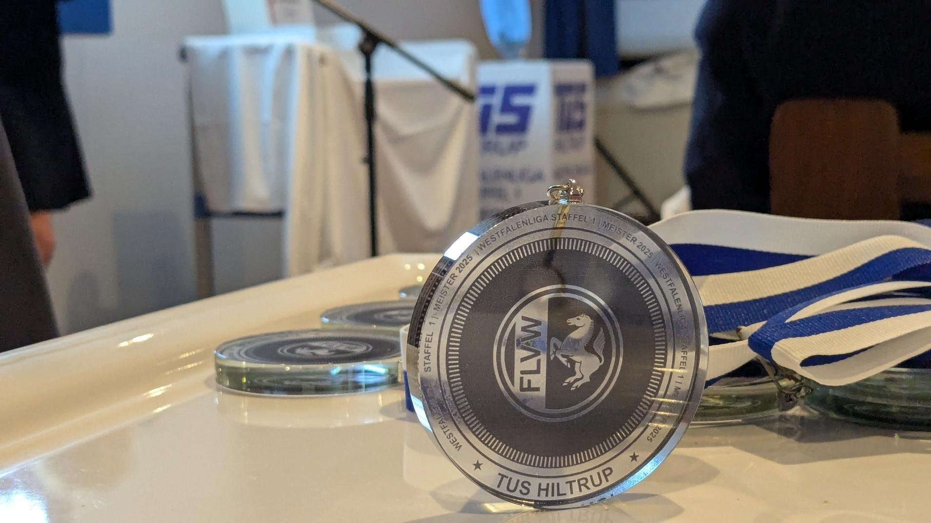 Nahaufnahme einer Siegermedaille mit der Gravur „TUS Hiltrup“ auf einem Tisch, umgeben von weiteren Medaillen.