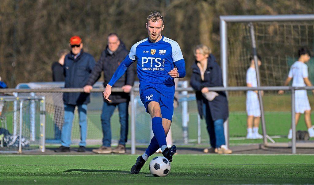 Ein Fußballspieler in blauer Uniform dribbelt mit einem Ball auf einem Kunstrasenplatz, Zuschauer im Hintergrund.