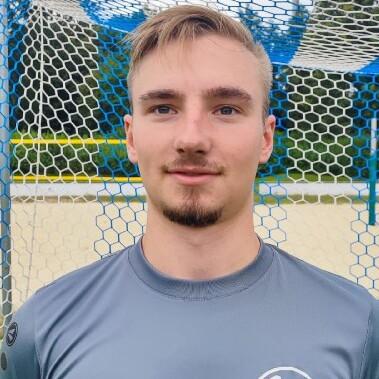 Junger Mann in grauer Sportbekleidung steht vor einem Fußballtor im Freien, mit einem Netz im Hintergrund.