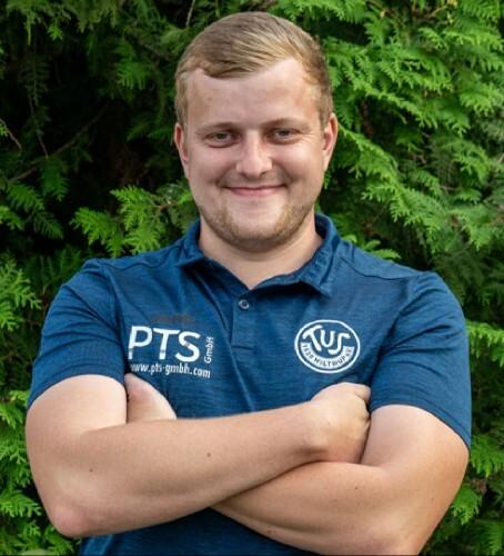 Lächelnder junger Mann mit verschränkten Armen, trägt ein blaues Polo-Shirt vor einer grünen Hintergrundlandschaft.