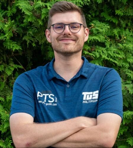 Mann mit Brille und verschränkten Armen in blauer Poloshirt vor grünem Hintergrund.