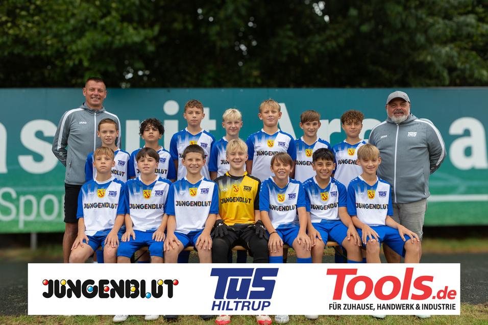 Mannschaftsfoto einer Jugendfußballmannschaft mit Trainer, in blauen Trikots auf einem Sportplatz.