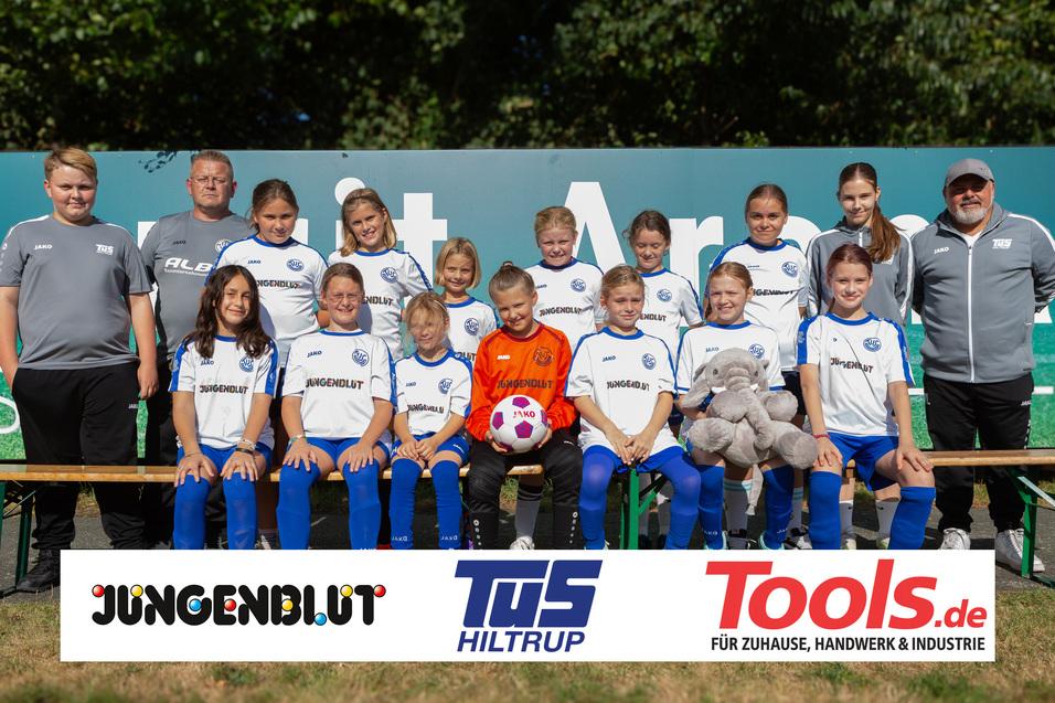 Mädchenfußballmannschaft mit Trainer, in weißen Trikots, Positur vor einem grünen Banner und einem Fußball.