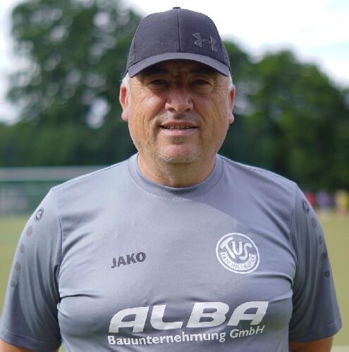 Mann mit Cap steht lächelnd vor einem Fußballplatz, trägt graues Teamtrikot mit Logo und Sponsorenaufdruck.