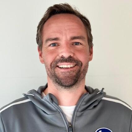 Porträt eines lächelnden Mannes mit kurzem Haar, in grauer Sportjacke mit Logo und Aufschrift „JAKO“.