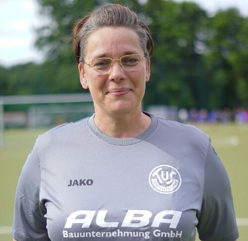 Portrait einer Frau in grauem Sporttrikot, lächelnd, mit Brille und Stadionhintergrund.