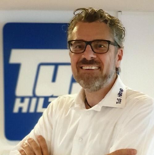 Lächelnder Mann mit Brille, weiße Bluse, vor einem Hintergrund mit Logo von TUS Hilft.