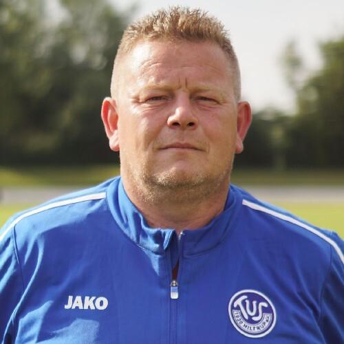 Porträt eines Trainers in blauer Sportjacke, mit ernstem Ausdruck, vor unscharfem Hintergrund.