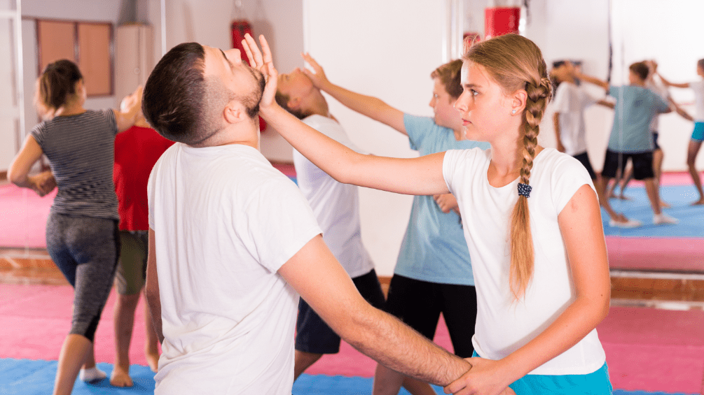 Kurs Rolling Selfdefense Zwei Personen üben Selbstverteidigung in einem Trainingsraum mit Matten und Spiegeln, weitere Teilnehmer im Hintergrund.