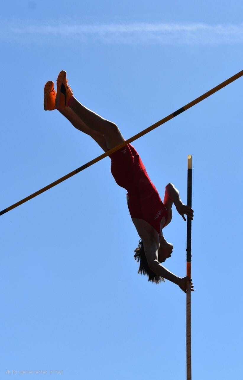 Sportlerin beim Stabhochsprung, kopfüber über eine Stange vor blauem Himmel, in rotem Outfit und orangefarbenen Schuhen.