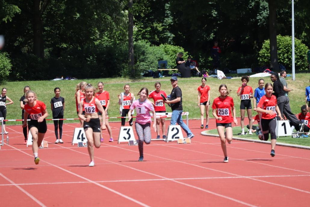 Sechs junge Athletinnen bei einem Wettlauf auf einer roten Tartanbahn, Zuschauer im Hintergrund.