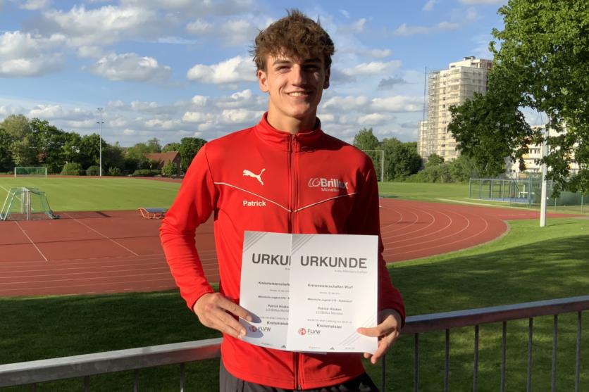 Sportler in roter Jacke hält Urkunden auf einem Sportplatz mit Rasen und Laufbahn im Hintergrund.