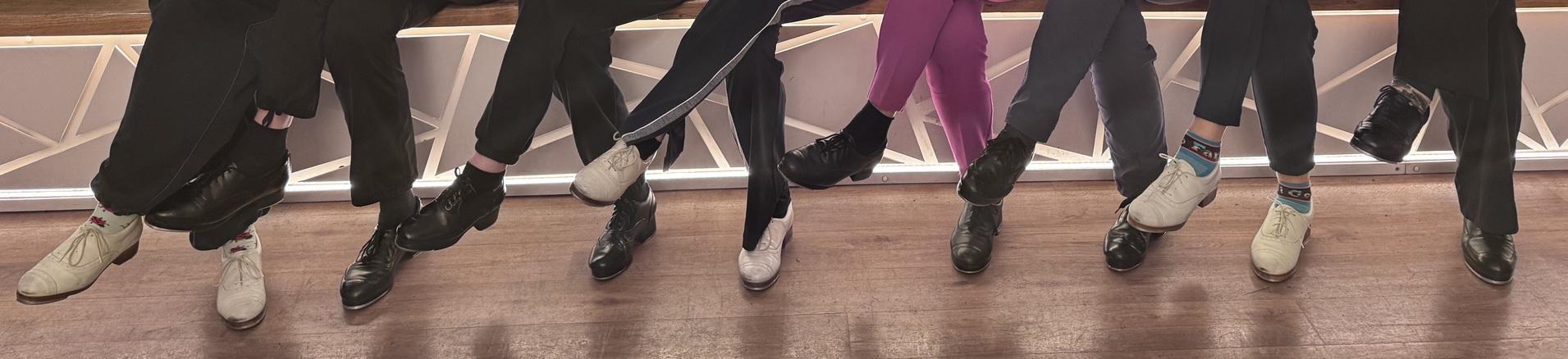 Reihe von Füßen in verschiedenen Schuhen, die über einer Holzoberfläche hängen, teils mit bunten Socken.