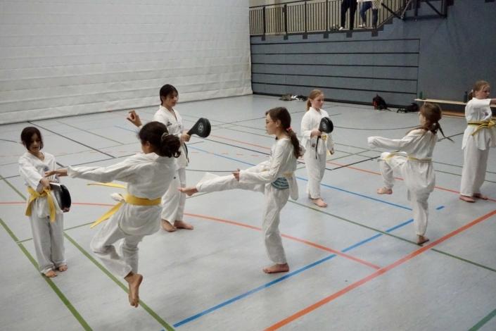 Sechs Kinder in weißen Karateanzügen üben in einer Sporthalle mit Gelbe-Gürtel-Techniken.