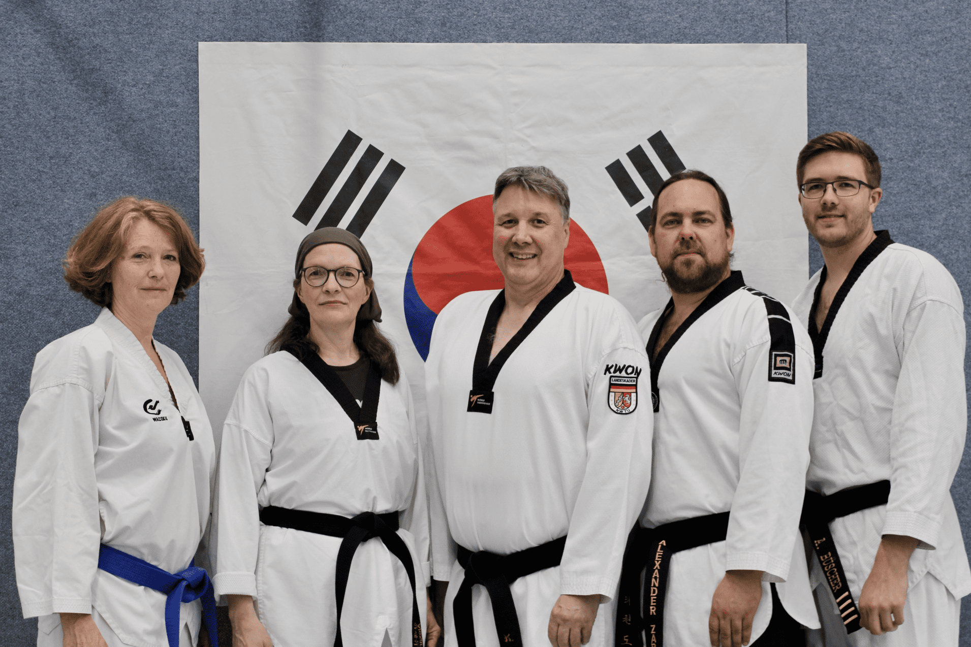 Fünf Taekwondo-Praktizierende in Trainingsanzügen stehen vor einer Flagge Südkoreas.