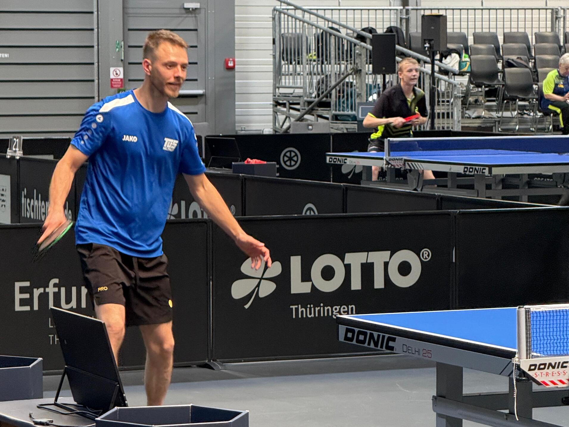 Mann im blauen Trikot spielt Tischtennis, während ein anderer Spieler im Hintergrund sich vorbereitet. Tischtennisplatte sichtbar.