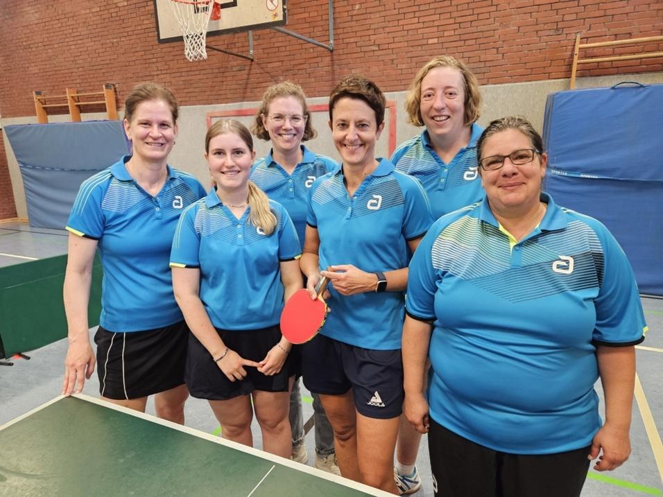Sechs Frauen in blauen Tischtennistrikots posieren lächelnd vor einem Tischtennistisch in einer Sporthalle.