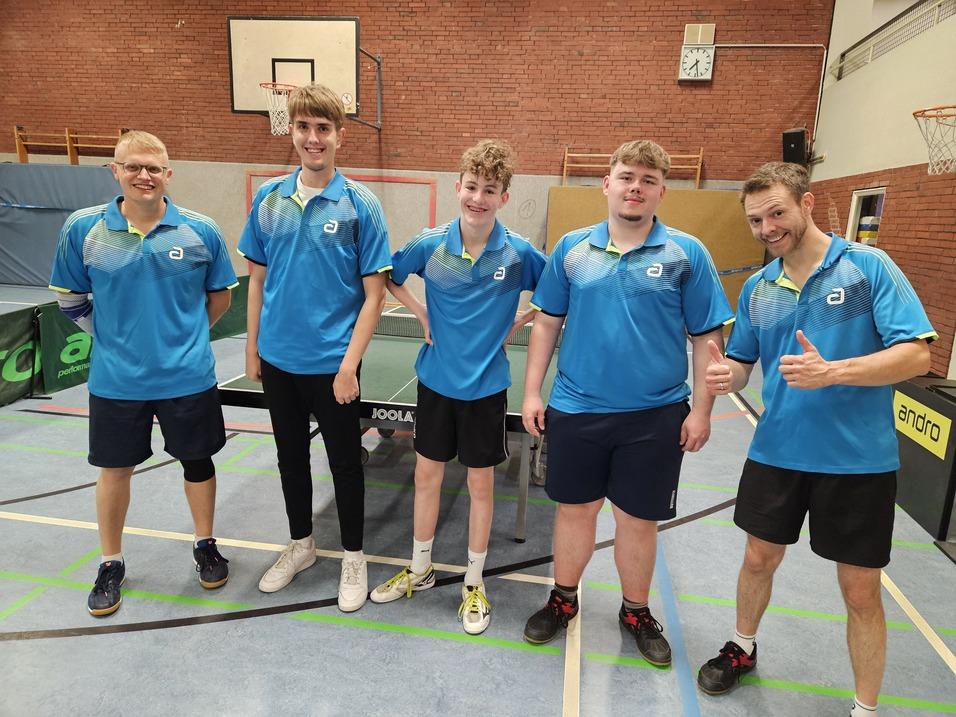 Fünf Tischtennisspieler in blauen Trikots stehen lächelnd vor einem Tischtennistisch in einer Sporthalle.