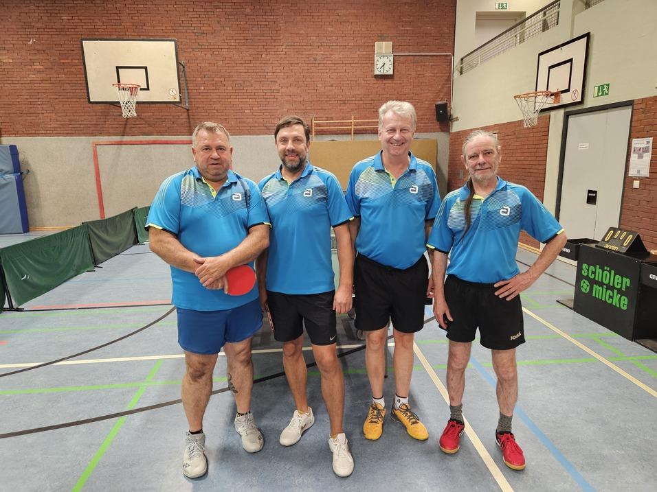 Vier Männer in blauen Tischtennistrikots stehen in einer Sporthalle mit Tischtennisplatte und Basketballkorb im Hintergrund.
