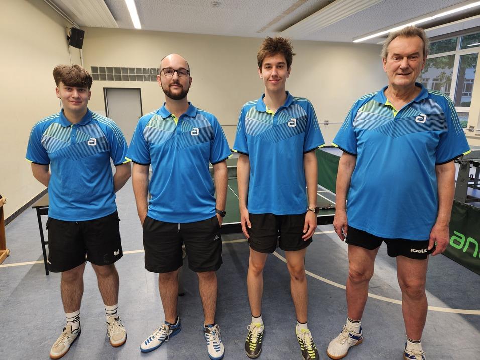 Vier Tischtennisspieler in blauen Trikots stehen vor einer Tischtennisplatte in einer Sporthalle.
