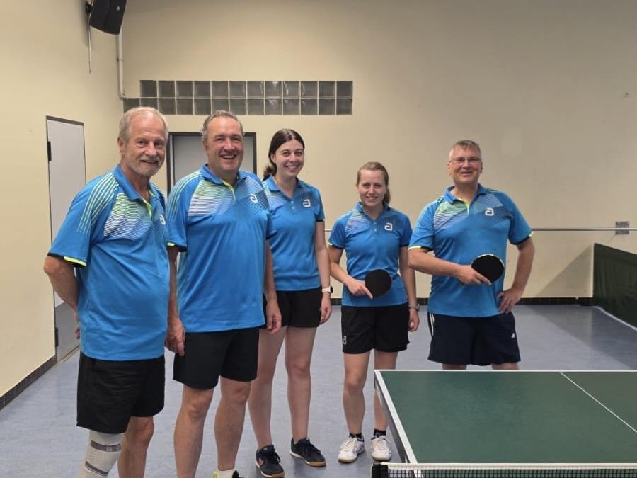 Fünf Personen in blauen Tischtenniskleidern stehen lächelnd neben einem Tischtennistisch in einer Sporthalle.