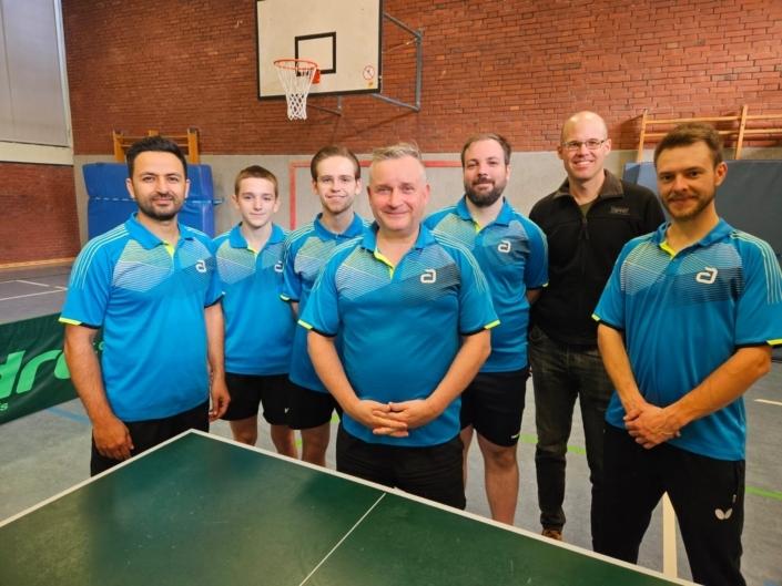 Gruppe von sechs Tischtennisspielern in blauen Trikots steht vor einem Tischtennistisch in einer Sporthalle.