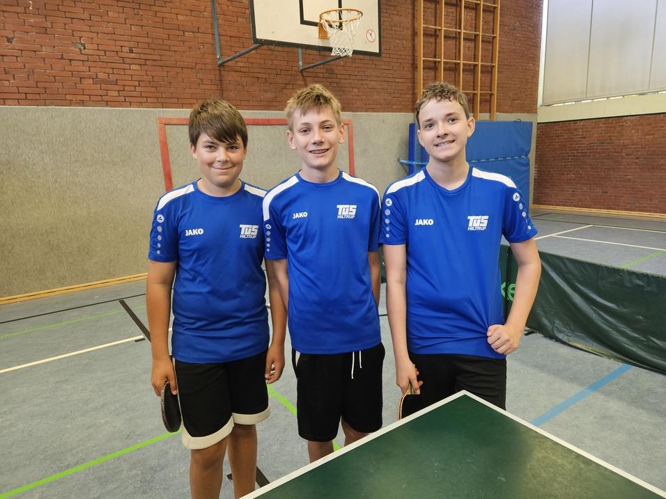 Drei Jungen in blauen Sporttrikots stehen lächelnd vor einem Tischtennistisch in einer Sporthalle.