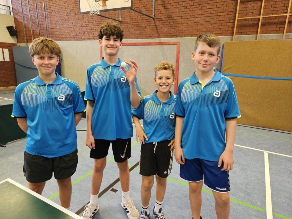 Vier Jungen in blauen Sporthemden stehen lächelnd zusammen in einer Turnhalle, der eine hält einen Tischtennisball in der Hand.