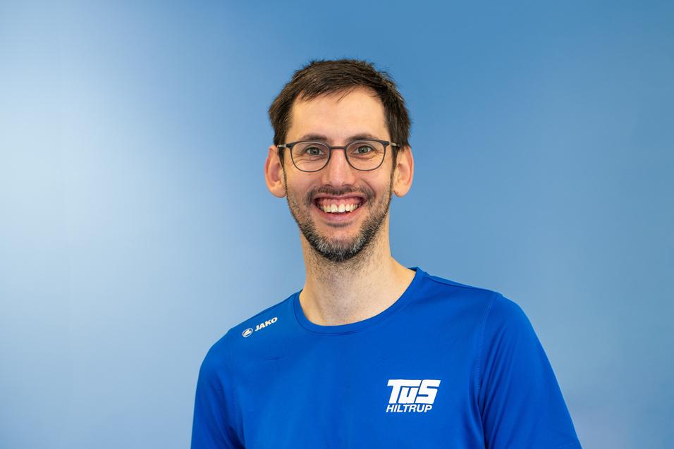 Lächelnder Mann mit Brille trägt ein blaues Sport-T-Shirt vor einem hellblauen Hintergrund.