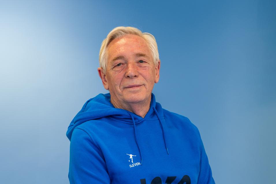 Älterer Mann mit kurzen grauen Haaren, trägt einen blauen Kapuzenpullover, vor einfarbig blauer Wand.