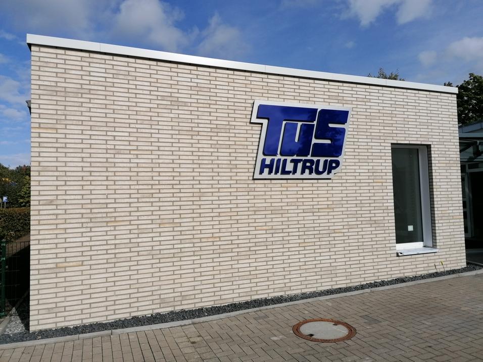 Fassade eines Gebäudes mit dem blauen Schriftzug „TuS Hiltrup“ auf hellen Ziegeln und einem Fenster.