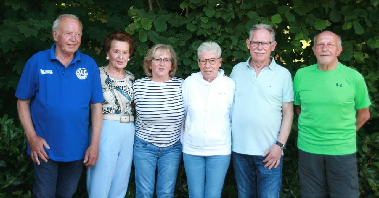Gruppenfoto von sechs Personen vor einem grünen Hintergrund, lächelnd und teilweise Schulter an Schulter stehend.