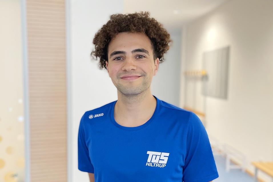 Chedi Touati Junger Mann mit lockigem Haar in blauem Sportshirt lächelt vor einem hellen, modernen Hintergrund.