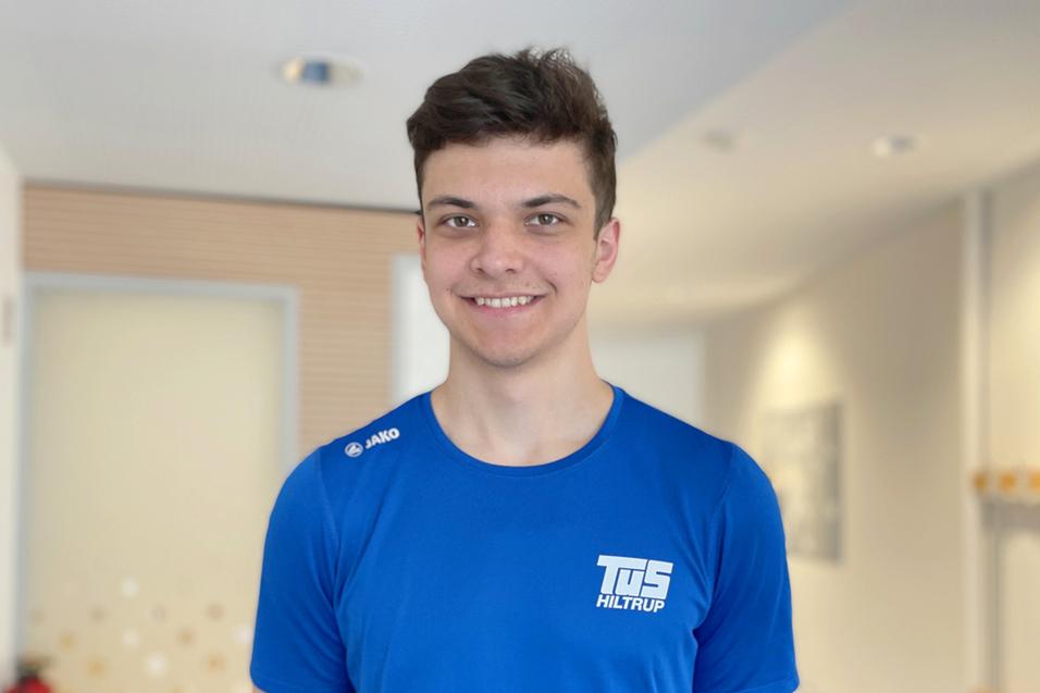 Emmanuel Dudzik Lächelnder junger Mann trägt ein blaues T-Shirt mit dem Schriftzug „TuS Hiltrup“, Hintergrund unscharf mit hellem Raum.