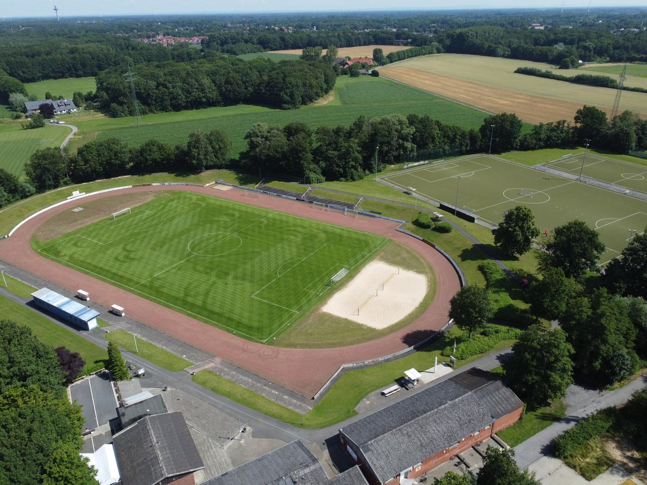Luftaufnahme eines Fußballfeldes mit Leichtathletikbahn, Sandkasten und angrenzenden Sportplätzen umgeben von Wald und Feldern.