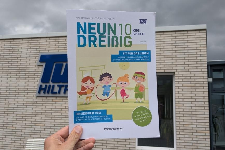 Hand hält ein Vereinsmagazin des TuS Hiltrup mit dem Titel „NEUN DREIßIG KIDS SPECIAL“ vor einer Gebäude-Fassade.