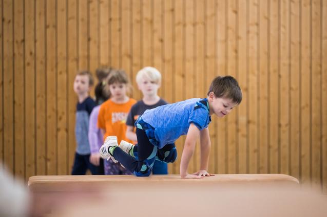 Ein Junge balanciert auf einem Blickkasten, während mehrere Kinder im Hintergrund zuschauen. Holzpaneele sind sichtbar.